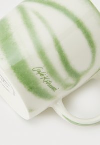 Κεραμική κούπα με λευκό σώμα και πράσινα μοτίβα νερομπογιάς σε σχήμα σπείρας. Φέρει την επιγραφή "Café Kitsuné" με πράσινο κείμενο. Κυρτή λαβή, λεία υφή.