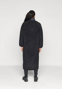 Zwarte textuur lange jas met een losse silhouet en lange mouwen, voorzien van een kraag en reikt tot halverwege de kuit. Draagt over zwarte broek en schoenen.