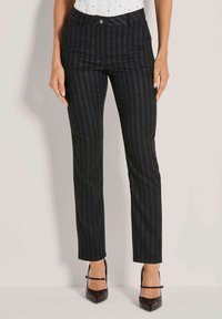 Vrouw in donkergrijze verticaal gestreepte broek en zwarte puntige hakken met enkelbandjes, staande tegen een effen lichte achtergrond.