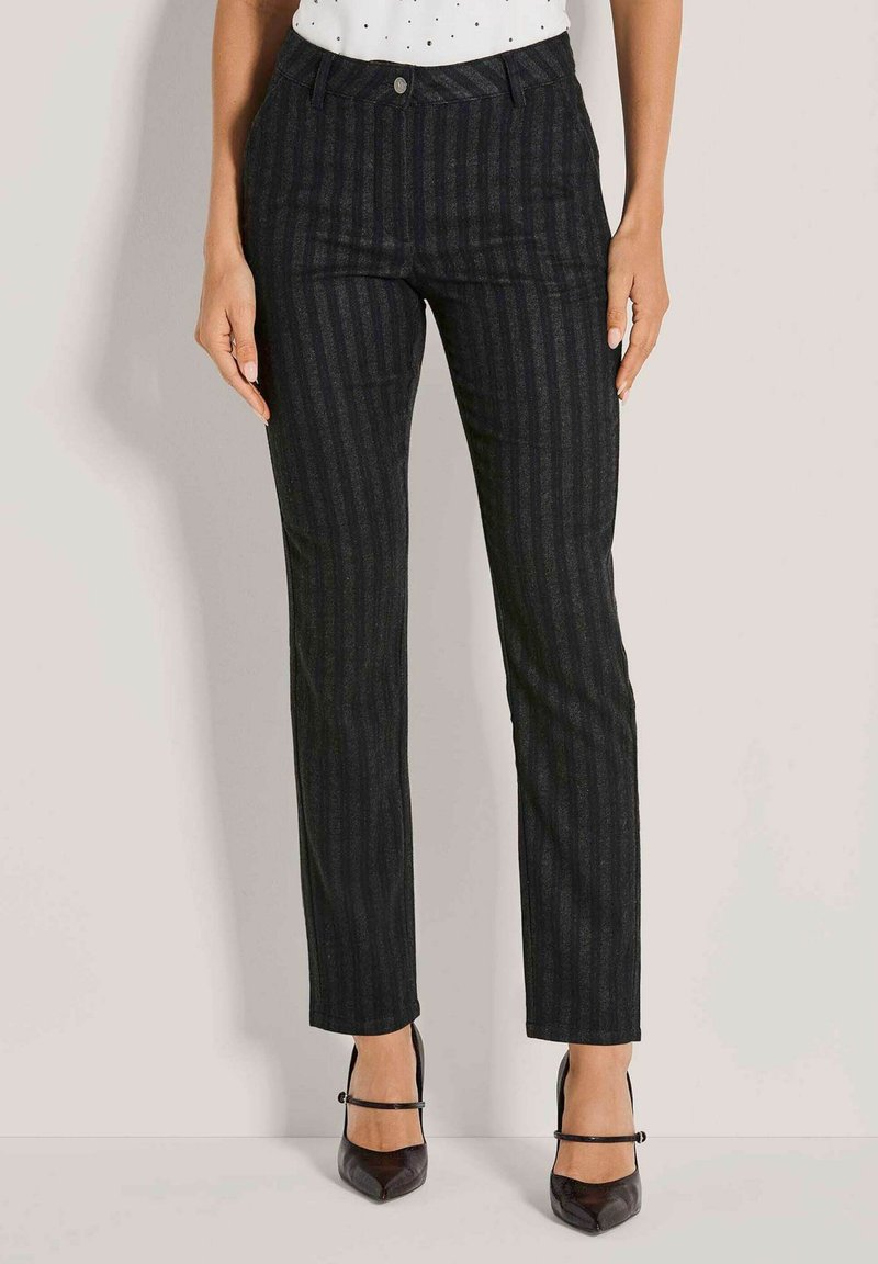 Vrouw in donkergrijze verticaal gestreepte broek en zwarte puntige hakken met enkelbandjes, staande tegen een effen lichte achtergrond.