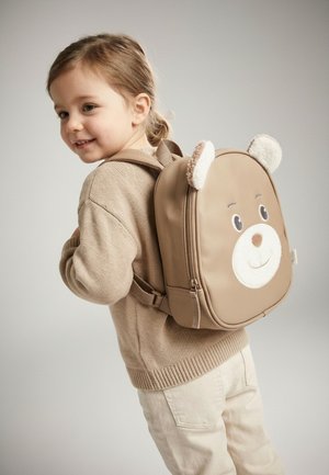 Young child smiling, wearing beige sweater and pants, with a tan backpack featuring a bear face and soft ears.