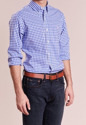Man draagt een blauw-wit geruit overhemd met opgerolde mouwen, een bruine riem en donkerblauwe jeans.