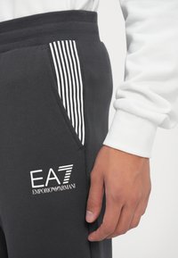 Tuva plāksne ar roku blakus melnajām sporta biksēm, uz kurām ir baltais EA7 Emporio Armani logo un svītrainās kabatas detaļa, apvienotas ar baltu garām piedurknēm topu.