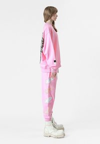 Sudadera rosa con diseño de alas en la parte posterior, combinada con pantalones de chándal rosas a juego que presentan alas bordadas en blanco y rosa. Botas blancas.