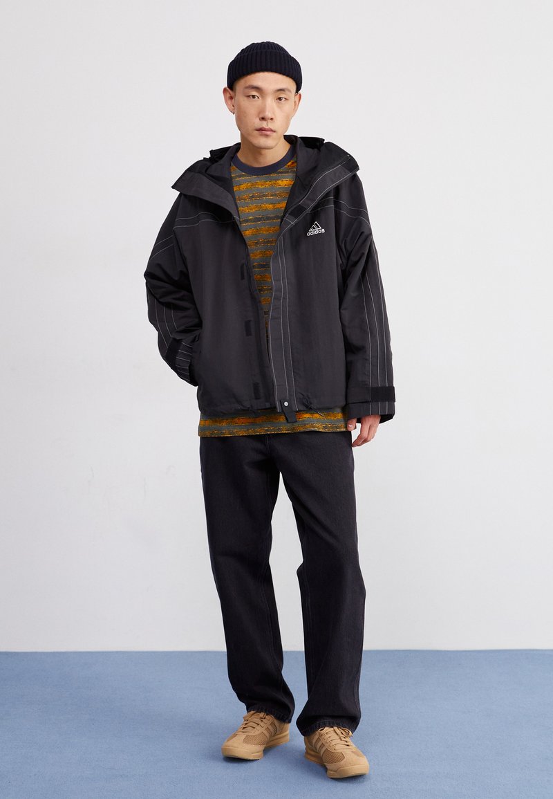 Chaqueta negra impermeable con costuras blancas, botones frontales y capucha. Debajo, una camiseta rayada amarilla y gris, combinada con jeans oscuros y sneakers color beige.