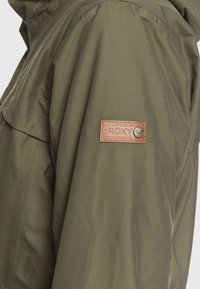 Manche de veste vert olive avec écusson marron Roxy cousu et petit emblème métallique près de l'épaule.