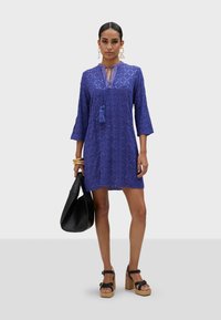 Blå klänning med trekvartsärmar, med ett diamantsmönster och fransdetalj vid halslinningen. Bärs med svart väska och platåsandaler.