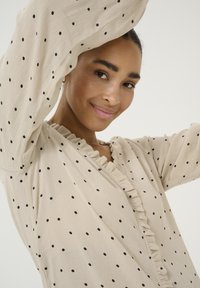 Beige blouse with black polka dots, ruffled V-neckline, and long sleeves. Lightweight, textured fabric.