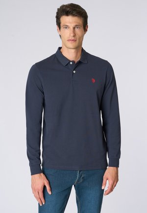 Uomo che indossa una polo a maniche lunghe blu navy con un logo rosso ricamato sul petto e jeans blu, in piedi davanti a uno sfondo neutro.