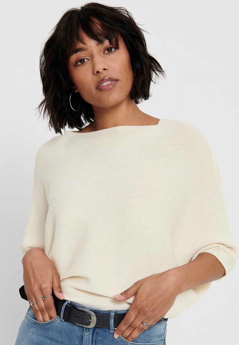 Cream, ribbed knit sweater with a boat neckline and loose fit. Paired with blue jeans and a black belt, featuring silver buckle and rings.