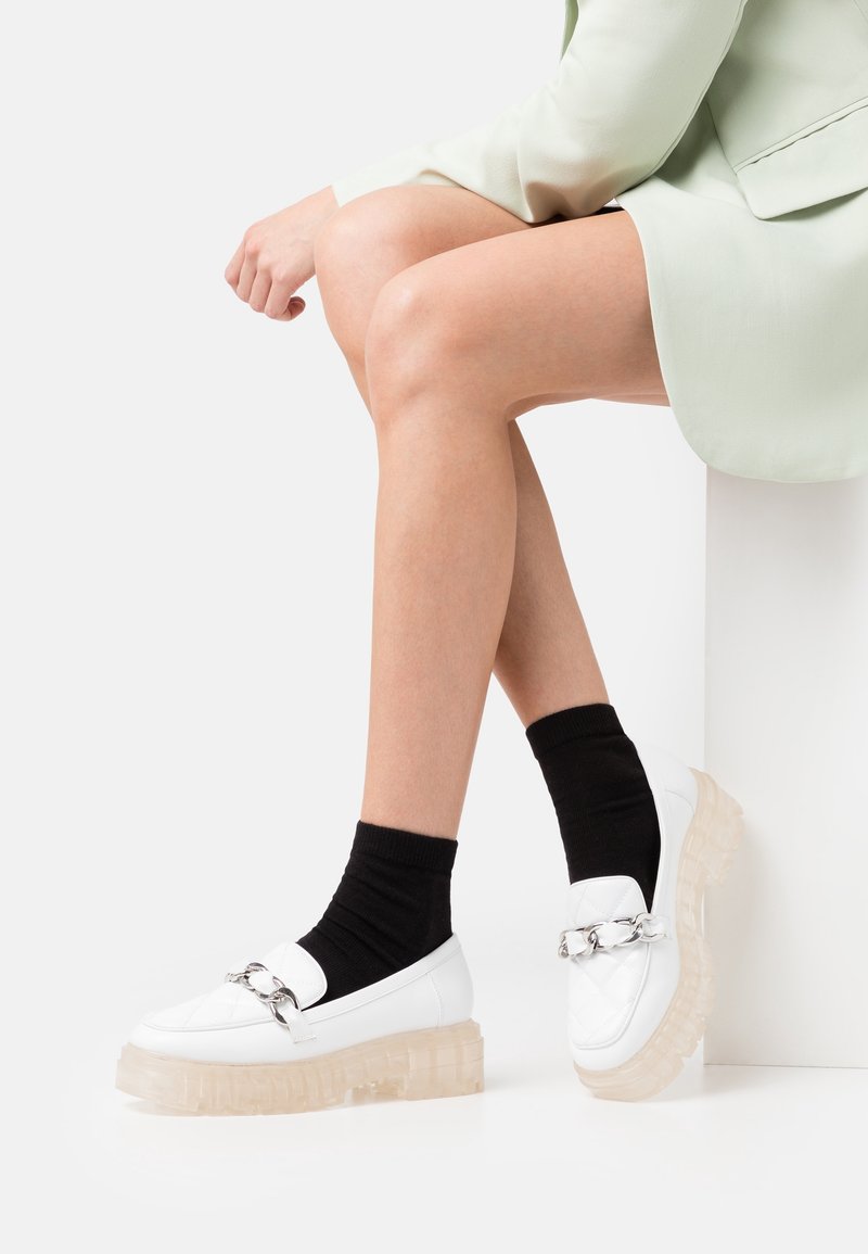 Mocassins blancs matelassés avec une semelle épaisse transparente et un détail en chaîne argentée, associés à des chaussettes noires à mi-mollet. La chaussure a une forme arrondie.