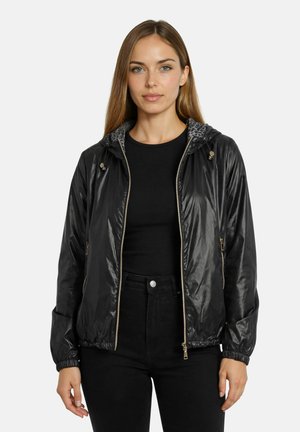 Femme aux cheveux lisses châtain clair portant une veste noire brillante zippée par-dessus une chemise noire et un pantalon noir sur fond blanc.