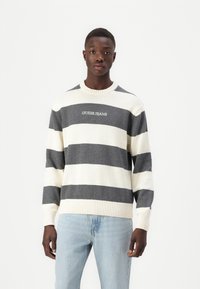 Csíkos pulóver fehér és sötétszürke színben, bordázott mandzsettákkal és alsó szegéllyel, a mellkas közepén a "GUESS JEANS" logóval.