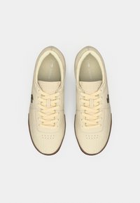 Μπεζ αθλητικά παπούτσια με υφασμένο επάνω μέρος, κρέμ κορδόνια και λαστιχένια σόλα. Διαθέτουν μικρό σκούρο λογότυπο στο πλάι και διάτρητες λεπτομέρειες.