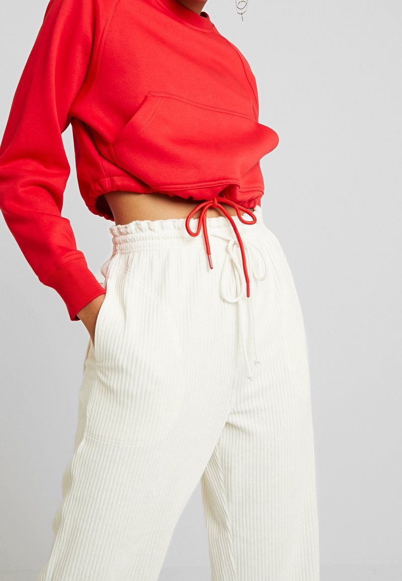 Personne portant un top court rouge à manches longues avec cordon et un pantalon blanc côtelé taille haute avec poches avant.