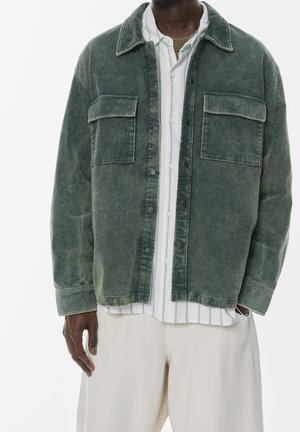 Homme portant une veste en velours côtelé verte sur une chemise blanche à rayures boutonnée et un pantalon beige clair, les mains détendues le long du corps.