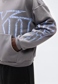 Person trägt einen grauen Pullover mit blauen, rissigen grafischen Designs, die Hand in der vorderen Tasche, und schwarze Hose vor einem einfarbigen Hintergrund.