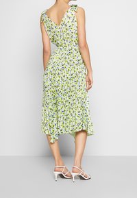 Frau in einem ärmellosen, V-ausschnitt, plissierten Midikleid mit grünem, blauem und weißem Blumenmuster, mit gebundenen Schulterträgern und weißen Absatzsandalen.