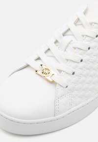 Baskets blanches avec un panneau latéral texturé en tissu tissé, des lacets blancs et une étiquette logo en métal doré sur les lacets.