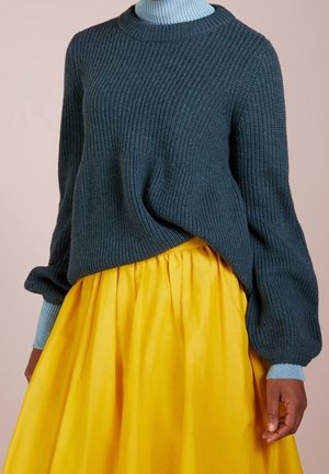 Chunky mørk teal strikket sweater med ribbet tekstur og lange, puffede ærmer. Parret med en lys gul plisseret nederdel.