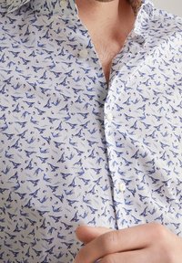 Wit shirt met een patroon van blauwe vogels, een korte kraag en een knoopsluiting aan de voorkant. Zachte stof met een subtiele textuur.