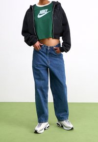 Svart croppad hoodie över en grön Nike-t-shirt, kombinerad med högmidjade blå denimjeans och vita sneakers med svarta detaljer.