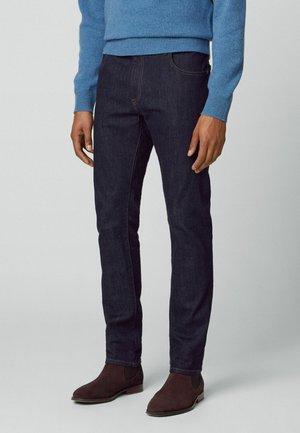 Mørkeblå jeans med slim fit, der har fem lommer og subtile syninger, parret med en lyseblå striktrøje og brune støvler.