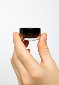 Brun gelprodukt i en klar burk med svart lock, hålls av en hand. Märkesnamnet "INGLOT" synligt på burkens sida. Slät textur.