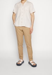 Kortärmad beige skjorta med bröstfickor, ljusbruna slim-fit byxor och vita sandaler med mörk sula och spännedetalj.