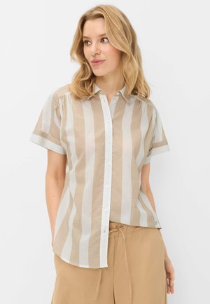 Femme aux cheveux blonds porte une chemise à manches courtes boutonnée à rayures verticales beige et blanche et un pantalon beige avec cordon, regardant sur le côté.