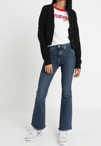 Černý žebrovaný kardigan přes bílý tričko s červeným textem, spárovaný s modrými rozšířenými džínami a bílými teniskami s černými akcenty.