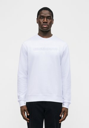 Ung mand med mørk hud iført en hvid sweatshirt med præget tekst "Versace Jeans Couture" og sorte bukser, stående foran en ensfarvet baggrund.