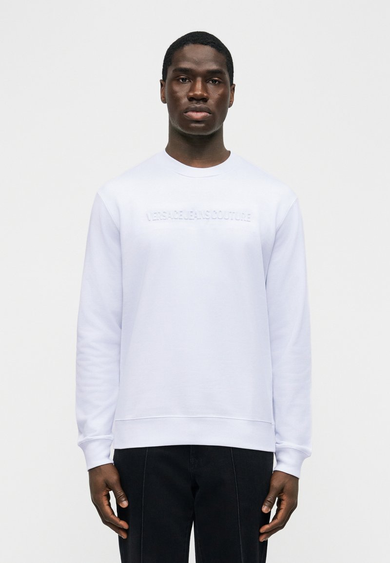 Młody mężczyzna o ciemnej karnacji w białej bluzie z wypukłym napisem "Versace Jeans Couture" i czarnych spodniach, stojący na gładkim tle.