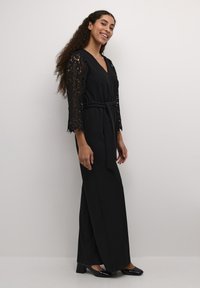 Tuta nera con intricate maniche in pizzo a tre quarti, scollatura a V e una cintura in tessuto in vita; gambe dell pantaloni svasate e eleganti.