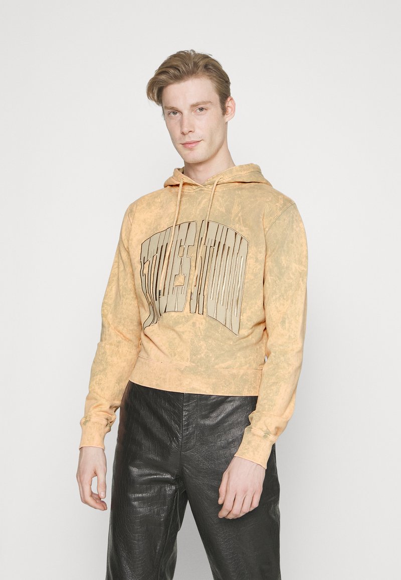 Κοντομάνικη hoodie με σχέδιο tie-dye σε ανοιχτό πορτοκαλί χρώμα, με μεγάλο, ανάγλυφο γραφικό μπροστά. Μαλακό ύφασμα και ριμπ μανσέτες, συνδυασμένο με σκούρες δερμάτινες παντελόνες.