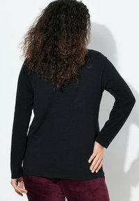 Schwarzes Langarmshirt aus weichem Stoff, mit einem lockeren Schnitt und rundem Halsausschnitt. Das Modell trägt es mit burgunderfarbenen Hosen.