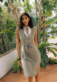 Femme aux cheveux foncés mi-longs portant une robe sans manches boutonnée et un foulard autour du cou, debout à l'extérieur parmi des plantes tropicales.