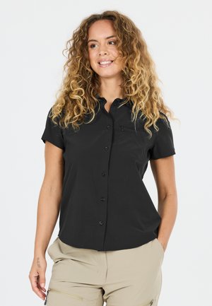 Frau mit lockigem Haar, die ein schwarzes kurzärmeliges Hemd mit Knöpfen und beige Hosen trägt, lächelt und posiert mit den Händen in den Taschen.