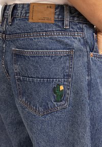 Blauwe denim jeans met een leren label op de tailleband en een cactus borduursel detail op de achterzak.
