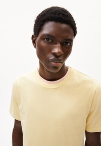 Helles gelbes T-Shirt aus Baumwolle mit rundem Halsausschnitt und kurzen Ärmeln. Auffälliges Detail am inneren Kragen in Pink. Glatte Textur.