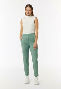 Weißes ärmelloses Rollkragenoberteil mit strukturiertem Muster, kombiniert mit mintgrünen maßgeschneiderten Hosen und weißen Sneakers.
