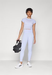 Svetlofialový crop top a zodpovedajúce vysoké legíny, vyrobené z elastického materiálu. Čierna športová taška s bielym logom, biele tenisky so pruhmi.