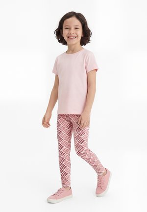 Dievča s kučeravými vlasmi, usmievajúce sa, kráčajúce, oblečené v svetloružovom tričku, vzorovaných ružových legínach a ružových teniskách na bielom pozadí.