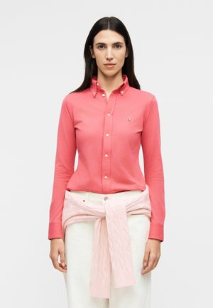 Γυναίκα με μακριά σκουρόχρωμα μαλλιά, φορώντας ένα κοραλί πουκάμισο με κουμπιά και ένα ανοιχτό ροζ πουλόβερ δεμένο στη μέση της πάνω από λευκό παντελόνι.