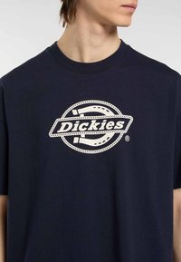 Námořnická bavlněná tričko s bílým kruhovým logem, na kterém je v středovém designu text "Dickies" v podobě stuhy. Krátké rukávy, kulatý výstřih.