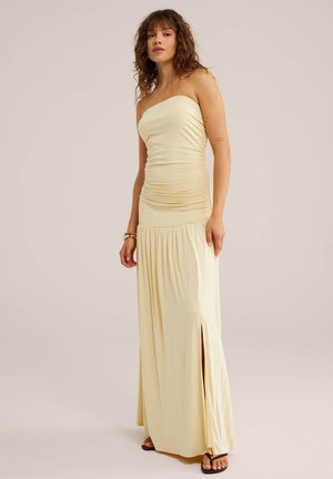 Žena s vlnitými hnedými vlasmi oblečená v nízkom vrchnom žltom dlhom šate s bočným rozparkom a hnedými sandálmi, stojí pred jednoduchým béžovým pozadím.