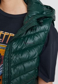 Osoba nosząca ciemnozieloną pikowaną kamizelkę bez rękawów na czarnym t-shircie z grafiką, na którym częściowo widoczne jest kolorowe napisy.