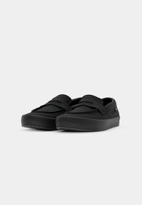 Sorte slip-on sko med flat, bred såle, mykt overmateriale og lagdelt design. Har en teksturert overflate og minimal merkevare.