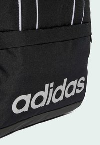 Mochila negra hecha de un tejido duradero, con rayas blancas y un destacado logo gris de "adidas" en la parte inferior. Textura suave, diseño deportivo.