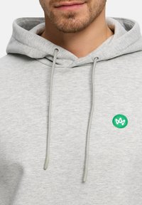 Man draagt een lichtgrijze hoodie met trekkoorden en een klein groen cirkelvormig logo op de borst met een wit abstract ontwerp.